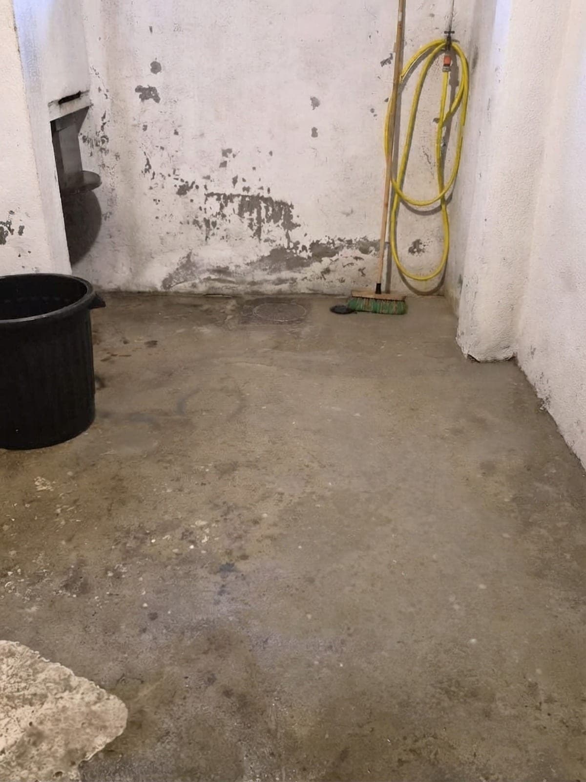 Cave au sol de béton nettoyé, murs blancs, tuyau jaune enroulé, balai et bac noir rangés